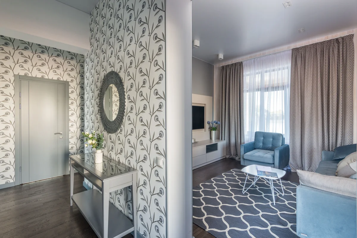 Tapezierarbeiten: Eingangsbereich mit präzise angebrachter, floral gemusterter Tapete, einem runden Spiegel und Blick ins modern renovierte Wohnzimmer.